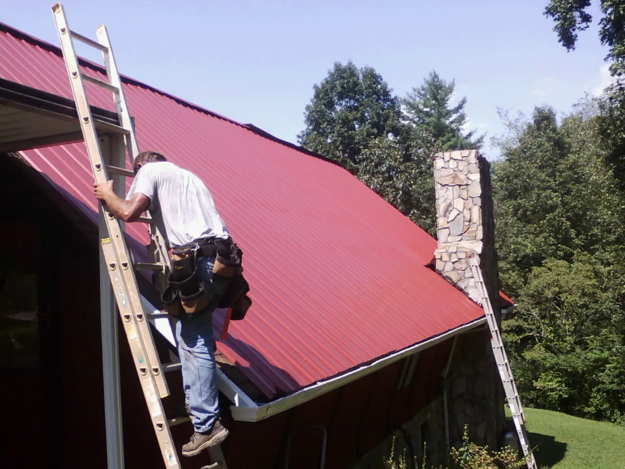 Roofer on a ladder beside a red metal roof and stone chimney — Soffit & Fascia Repair by Roofing Brothers in Deer Lodge