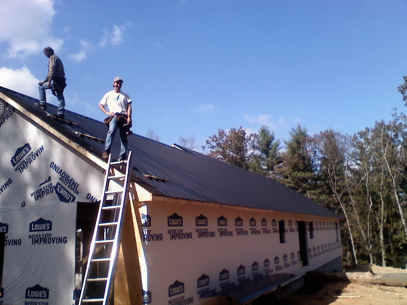 Two roofers standing on a long dark metal roof over a wrapped building in Deer Lodge